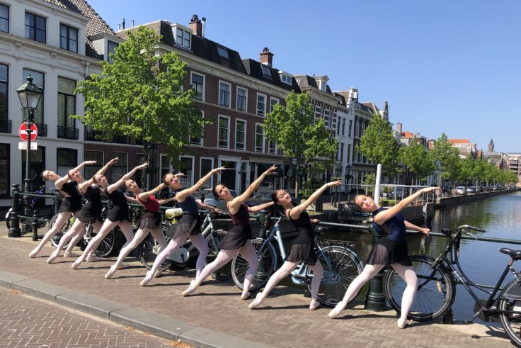 Students in ballet attire on a bridge over the canal near Toussaintkade and Noordwal in The Hague, practicing tendu with arm positions while using the railing like a barre at Summerschool Den Haag.